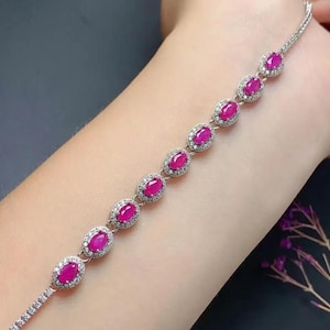 Puede incluir: Una pulsera de tono plateado que presenta una serie de piedras preciosas ovaladas de color rosa rojizo vibrante. Cada piedra está rodeada por un halo de pequeñas piedras claras y brillantes en un marco metálico, unidas por una fina cadena.