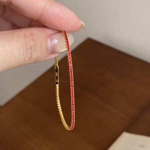 May include: A gold-colored bracelet with a line of small red gemstones. The bracelet is held up against a wooden surface and a book. The bracelet is a delicate piece of jewelry.