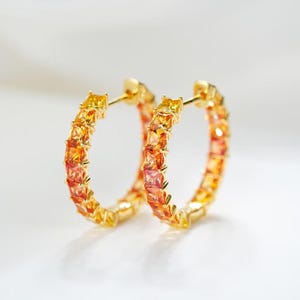 Könnte beinhalten: Ein Paar goldene Creolen mit einer durchgehenden Reihe quadratisch geschliffener Edelsteine in einem leuchtenden Farbverlauf von Gelb, Orange und Rot. Die Steine sind in einen Metallrahmen mit einem Klappverschluss eingefasst.