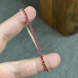 May include: A delicate tennis bracelet featuring a continuous row of small, round, vibrant pink-red gemstones set in a thin gold-toned metal chain. The bracelet is held straight, showcasing the uniform size and alignment of the stones.