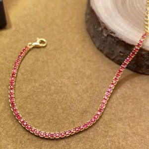 May include: A gold-colored bracelet with a delicate chain design. The bracelet is adorned with numerous small, vibrant red gemstones. The clasp is gold-colored. The bracelet is displayed on a neutral-toned surface.