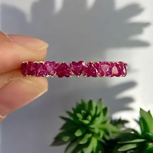 May include: A gold ring featuring a series of heart-shaped, deep red gemstones. The gemstones are set closely together, creating a continuous band of color. The ring is held against a soft background with a green plant in the lower right corner.