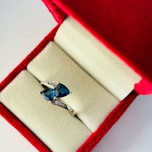 May include: A silver ring with two pear-shaped, deep blue gemstones and small clear stones. The ring is displayed in a red velvet jewelry box with a cream-colored interior.