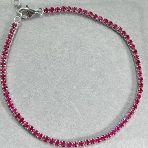 May include: A delicate silver bracelet featuring a continuous line of vibrant, round, ruby-red gemstones. The bracelet is set on a silver chain with a secure clasp. The gemstones are closely set, creating a sparkling, eye-catching piece of jewelry.