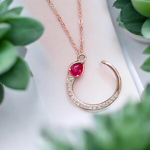 May include: Rose gold necklace featuring a teardrop-shaped, deep red gemstone pendant. The pendant is accented with small, clear crystals and is connected to a delicate chain. The necklace has a unique, open-circle design.
