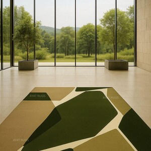 May include: A modern area rug with an abstract geometric design in shades of green, beige, and cream. The rug is rectangular and features the text "AYAT RUGS" in several places. The rug is placed on a tiled floor in front of a large window.