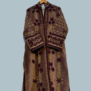 Pu&ograve; includere: Un lungo cappotto marrone con ricami dorati e bordeaux. Il cappotto presenta un collo alto e maniche lunghe, con motivi floreali e geometrici che adornano il davanti, le maniche e il colletto. &Egrave; appeso a una gruccia di legno.