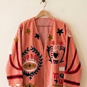 Puede incluir: Chaqueta color coral con diseños bordados. Presenta una parte delantera abierta y mangas largas. Los adornos incluyen un ojo, una mano, estrellas y otros símbolos en negro, blanco y dorado. La chaqueta cuelga de una percha de madera.