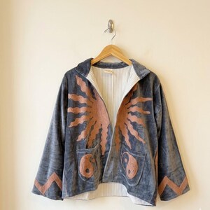 Puede incluir: Chaqueta de terciopelo gris con un diseño de sol en color óxido en la parte delantera y las mangas. La chaqueta tiene dos bolsillos con símbolos de yin y yang. El forro interior es a rayas. Colgada en una percha de madera.