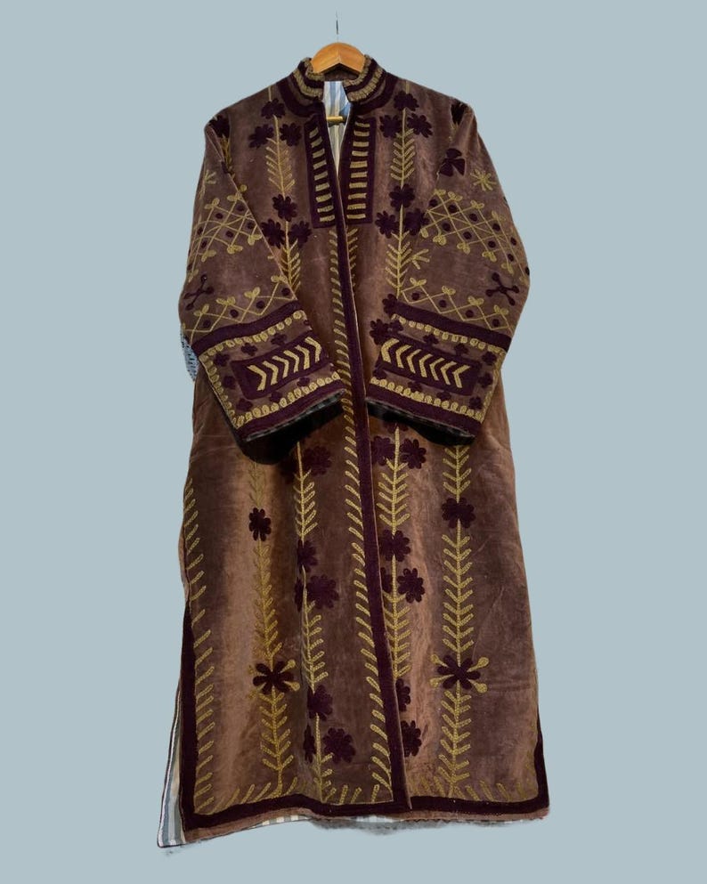 Pu&ograve; includere: Un lungo cappotto marrone con ricami dorati e bordeaux. Il cappotto presenta un collo alto e maniche lunghe, con motivi floreali e geometrici che adornano il davanti, le maniche e il colletto. &Egrave; appeso a una gruccia di legno.
