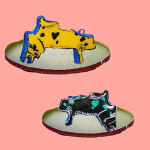Pode incluir: Dois ornamentos de vaca feitos à mão. Um é amarelo com manchas pretas e detalhes azuis, o outro é preto com manchas turquesa e detalhes azuis. Ambos estão sobre uma base redonda decorativa com uma borda vermelha e um centro verde claro. O fundo é rosa claro.
