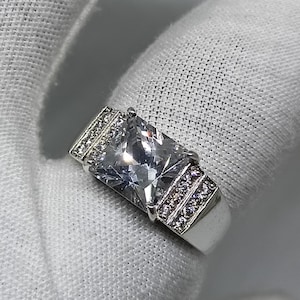 K&ouml;nnte beinhalten: Ein silberner Ring mit einem gro&szlig;en, rechteckigen, klaren Edelstein als Mittelpunkt. Das Band und die Seiten des Rings sind mit kleineren, klaren Edelsteinen verziert. Der Ring ist vor einem wei&szlig;en Hintergrund platziert.