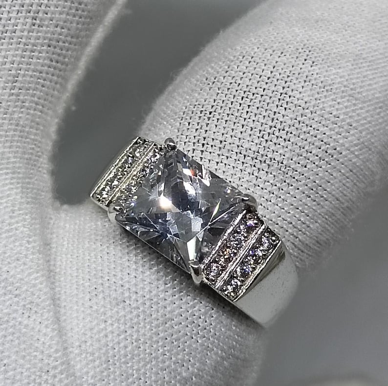 K&ouml;nnte beinhalten: Ein silberner Ring mit einem gro&szlig;en, rechteckigen, klaren Edelstein als Mittelpunkt. Das Band und die Seiten des Rings sind mit kleineren, klaren Edelsteinen verziert. Der Ring ist vor einem wei&szlig;en Hintergrund platziert.