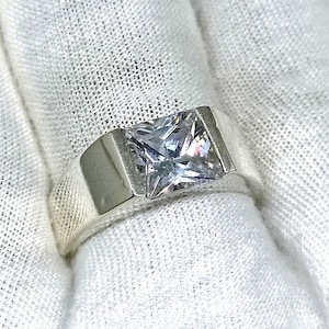 Può includere: Un anello d'argento con una grande gemma quadrata e trasparente. L'anello ha un design semplice e moderno con una fascia larga. La gemma è incastonata in una lunetta, su un tessuto bianco.