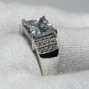 K&ouml;nnte beinhalten: Ein silberner Ring mit einem gro&szlig;en, quadratischen, klaren Edelstein als Mittelpunkt. Das Band hat eine Reihe kleinerer, klarer Steine. Der Ring ist vor einem wei&szlig;en Hintergrund platziert, der sein Funkeln und Design hervorhebt.