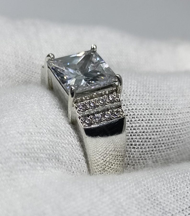K&ouml;nnte beinhalten: Ein silberner Ring mit einem gro&szlig;en, quadratischen, klaren Edelstein als Mittelpunkt. Das Band hat eine Reihe kleinerer, klarer Steine. Der Ring ist vor einem wei&szlig;en Hintergrund platziert, der sein Funkeln und Design hervorhebt.