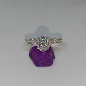 Op de afbeelding: Een zilveren verlovingsring met een grote ovaal geslepen diamant. De band heeft een gedraaid ontwerp met kleinere accentstenen. De ring wordt gepresenteerd op een klein, paars, afgerond object tegen een witte achtergrond.