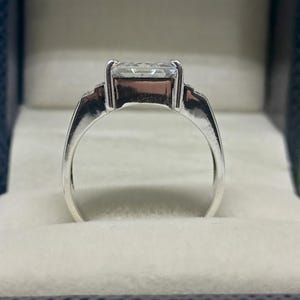K&ouml;nnte beinhalten: Ein silberner Ring mit einem gro&szlig;en, rechteckigen, klaren Edelstein. Der Ring hat ein schlichtes Band und ist in einer Vier-Zacken-Fassung gefasst. Der Ring wird in einer grauen Schmuckschatulle pr&auml;sentiert.
