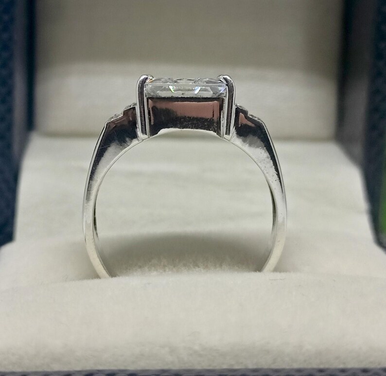 K&ouml;nnte beinhalten: Ein silberner Ring mit einem gro&szlig;en, rechteckigen, klaren Edelstein. Der Ring hat ein schlichtes Band und ist in einer Vier-Zacken-Fassung gefasst. Der Ring wird in einer grauen Schmuckschatulle pr&auml;sentiert.