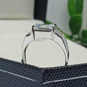 K&ouml;nnte beinhalten: Ein silberner Ring mit einem gro&szlig;en, rechteckigen, klaren Edelstein als Mittelpunkt. Der Ring hat einen schlichten Band und kleine Akzentsteine an den Seiten des Hauptsteins. Der Ring wird in einer schwarzen Schmuckschatulle pr&auml;sentiert.