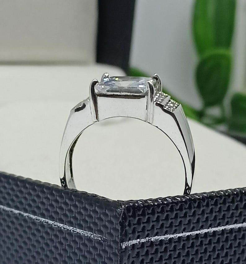 K&ouml;nnte beinhalten: Ein silberner Ring mit einem gro&szlig;en, rechteckigen, klaren Edelstein als Mittelpunkt. Der Ring hat einen schlichten Band und kleine Akzentsteine an den Seiten des Hauptsteins. Der Ring wird in einer schwarzen Schmuckschatulle pr&auml;sentiert.