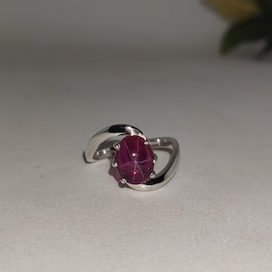 May include: A polished silver ring featuring a central oval-shaped cabochon star ruby. The deep magenta gemstone displays a distinct six-rayed star effect on its surface. The metal band curves around the stone, held in place by six small prongs.