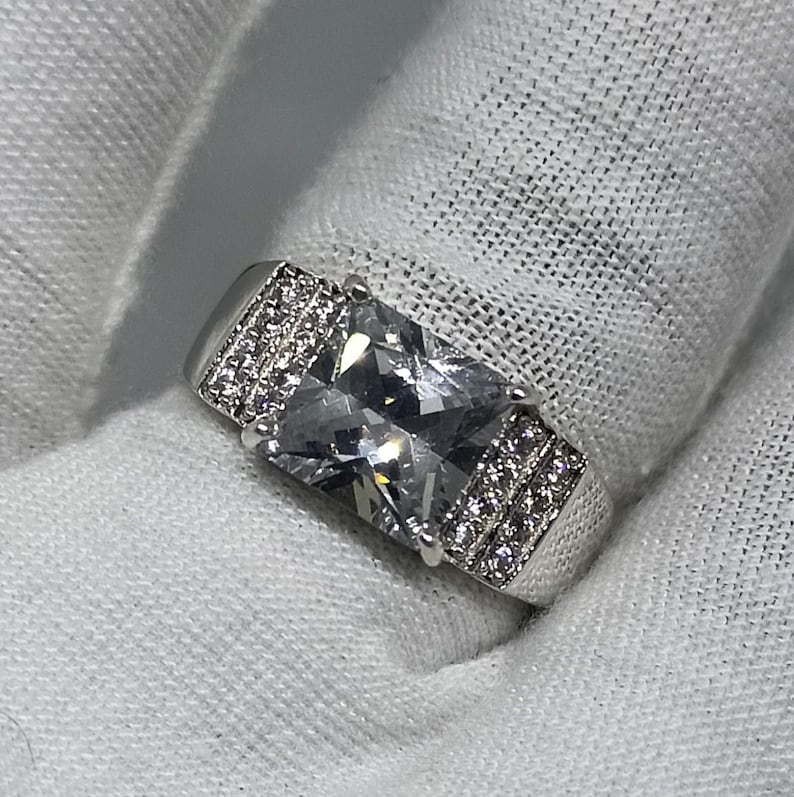 K&ouml;nnte beinhalten: Ein silberner Ring mit einem gro&szlig;en, rechteckigen, klaren Edelstein als Mittelpunkt. Das Band hat kleinere, klare Steine auf jeder Seite des Hauptsteins, die f&uuml;r Glanz sorgen. Der Ring ist vor einem wei&szlig;en Hintergrund platziert.