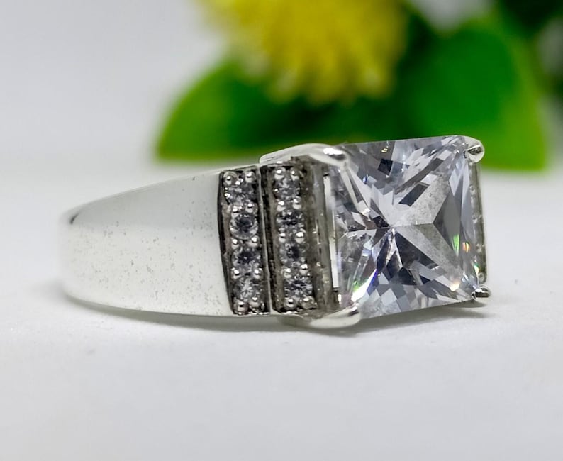 K&ouml;nnte beinhalten: Ein silberner Ring mit einem gro&szlig;en, quadratischen, klaren Edelstein. Das Band hat Reihen kleiner, klarer Steine auf beiden Seiten des Hauptsteins. Der Ring ist vor einem weichen, verschwommenen Hintergrund platziert.