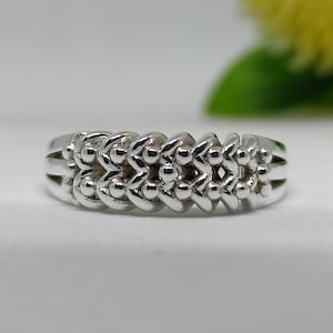May include: A silver ring with a repeating pattern of connected, rounded shapes. The ring has a polished finish and a delicate design, suitable for everyday wear. The ring is displayed on a white surface.