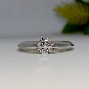 May include: A close-up of a silver ring with a large, clear, round-cut diamond. The diamond is the focal point, set in a minimalist, modern design. The ring band is smooth and polished, reflecting light. The background is blurred, highlighting the ring.