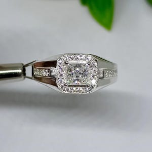 Op de afbeelding: Een zilveren ring met een vierkante, heldere edelsteen in het midden, omringd door kleinere, heldere stenen. Extra kleine stenen zijn langs de band van de ring gezet. De ring wordt vastgehouden door een zilveren gereedschap.