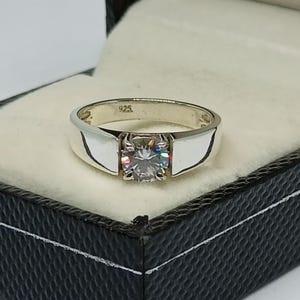 Può includere: Un anello d'argento con una grande gemma rotonda e trasparente incastonata in un'incastonatura a quattro griffe. La fascia è larga e lucida, con un design semplice e moderno. L'anello è presentato in un cofanetto nero.