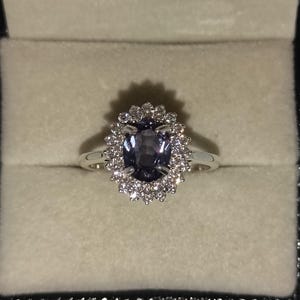 May include: A silver ring with a central oval, dark blue gemstone, encircled by a halo of smaller, clear stones. The ring is presented in a light-toned box, highlighting the dark blue gem.