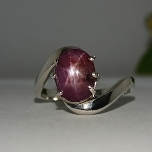 May include: A silver ring featuring a large, oval-shaped, deep red gemstone. The gemstone has a star-like effect. The ring's band has a flowing, curved design.