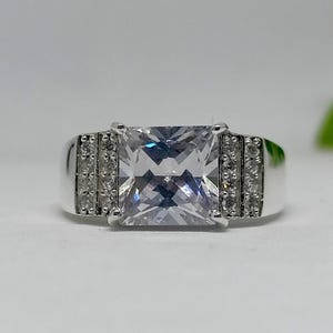 K&ouml;nnte beinhalten: Ein silberner Ring mit einem gro&szlig;en, quadratischen, klaren Edelstein als Mittelpunkt. Das Band hat kleinere, klare Steine auf beiden Seiten des Hauptsteins. Der Ring ist vor einem wei&szlig;en Hintergrund platziert.