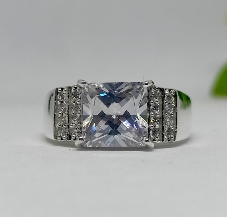K&ouml;nnte beinhalten: Ein silberner Ring mit einem gro&szlig;en, quadratischen, klaren Edelstein als Mittelpunkt. Das Band hat kleinere, klare Steine auf beiden Seiten des Hauptsteins. Der Ring ist vor einem wei&szlig;en Hintergrund platziert.
