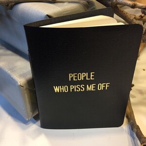May include: A black notebook with gold lettering that reads "PEOPLE WHO PISS ME OFF".