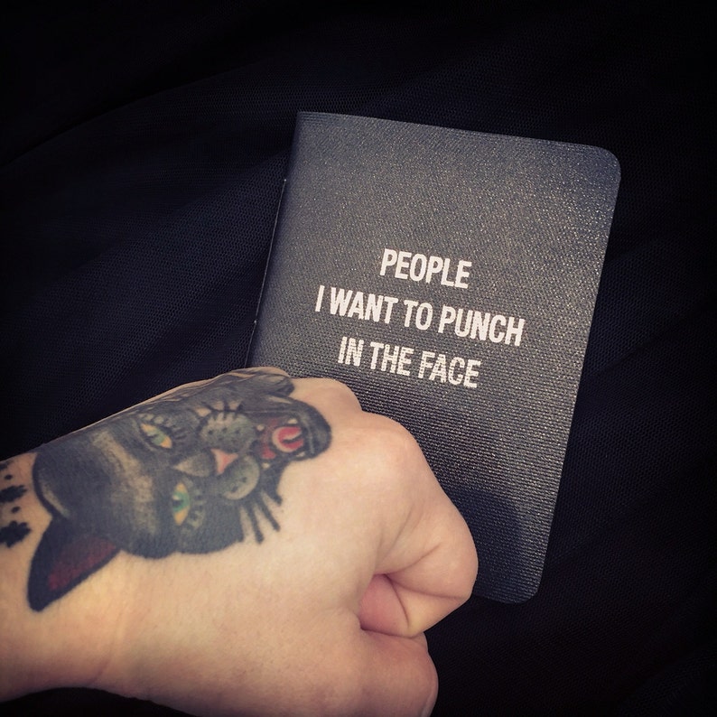 Może przedstawiać: Czarny notes z nadrukowanym białym tekstem "PEOPLE I WANT TO PUNCH IN THE FACE". Ręka z tatuażem czarnego kota trzyma notes.