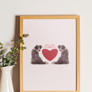 May include: A framed print featuring two dogs flanking a red heart with a pink bow. The dogs are depicted in shades of gray and black, with a light pink background. The frame is made of light brown wood.