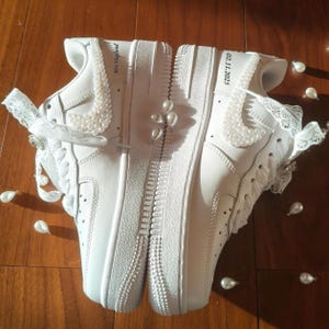 May include: White sneakers customized with pearl embellishments and lace accents. The Nike swoosh is adorned with pearls, and the text "Mrs Hoglund" and "02.11.2025" are visible. The shoes are set against a wooden surface, with scattered pearl-shaped decorations.