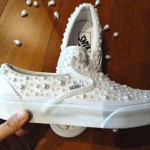 Könnte beinhalten: Weiße Slip-On-Sneaker, verziert mit zahlreichen Kunstperlen. Die Schuhe haben das Vans-Logo und stehen auf einer Holzoberfläche. Die Perlen sind um die Schuhe herum verstreut.
