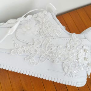 May include: A white sneaker featuring intricate white floral lace appliques stitched onto the side panel. The design includes delicate fabric flowers and small white pearl accents integrated into the lace pattern, paired with flat white satin ribbon laces.