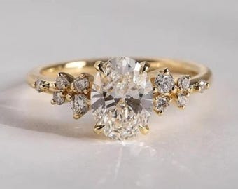 May include: A gold ring featuring an oval-cut diamond center stone. The ring is accented with smaller diamonds clustered on either side of the center stone. The band is a simple, polished gold. The ring is set against a white background.