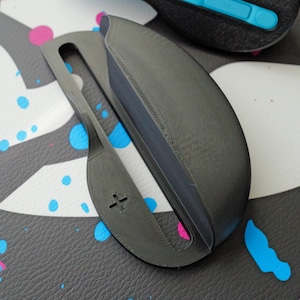 May include: A dark gray, 3D-printed object with a handle and a curved, enclosed section. The handle has a cross-shaped cutout. Another object with a blue accent is visible in the background. The objects are on a surface with a pattern of gray, white, pink, and blue paint splatters.