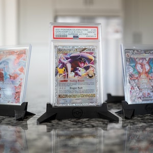 May include: Three Pokemon trading cards in protective cases, displayed on black stands. The center card features a Garchomp character with text that reads "Healing Breath" and "Dragon Rush". The cards are from the 2021 Pokemon Celebrations series.