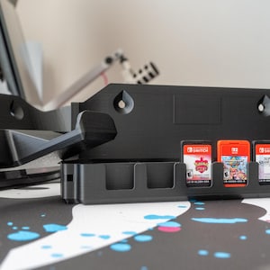 May include: A black 3D-printed game cartridge holder for Nintendo Switch games. The holder has slots for game cartridges and a larger section for the console. Three game cartridges are visible in the slots.