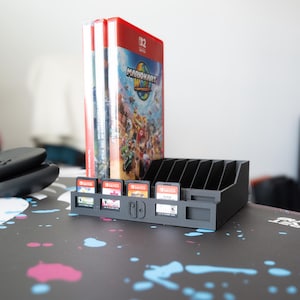 May include: A black Switch game cartridge holder with multiple slots, holding several game cartridges. Behind the holder are three game cases, with the Mario Kart 8 Deluxe case visible. A black controller is in the background.