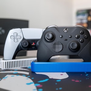 Puede incluir: Dos mandos de videojuegos, uno blanco y otro negro, descansan sobre soportes separados. El mando blanco tiene un logotipo de PlayStation, mientras que el mando negro tiene un logotipo de Xbox. Los soportes son blancos y azules, sobre una superficie estampada.