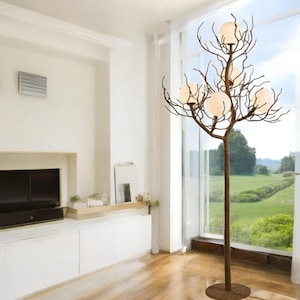 Puede incluir: Una lámpara de pie de color bronce con un diseño de ramas de árbol. La lámpara tiene seis pantallas esféricas de vidrio esmerilado. La lámpara se apoya en una base redonda y está situada frente a una gran ventana.