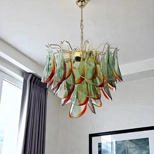 K&ouml;nnte beinhalten: Eine moderne H&auml;ngeleuchte mit goldfarbenem Rahmen und Kette. Die Leuchte verf&uuml;gt &uuml;ber zahlreiche glasf&ouml;rmige, bl&uuml;tenblattf&ouml;rmige Elemente in Gr&uuml;n-, Orange- und Klart&ouml;nen. Die Leuchte ist von der Decke abgeh&auml;ngt.